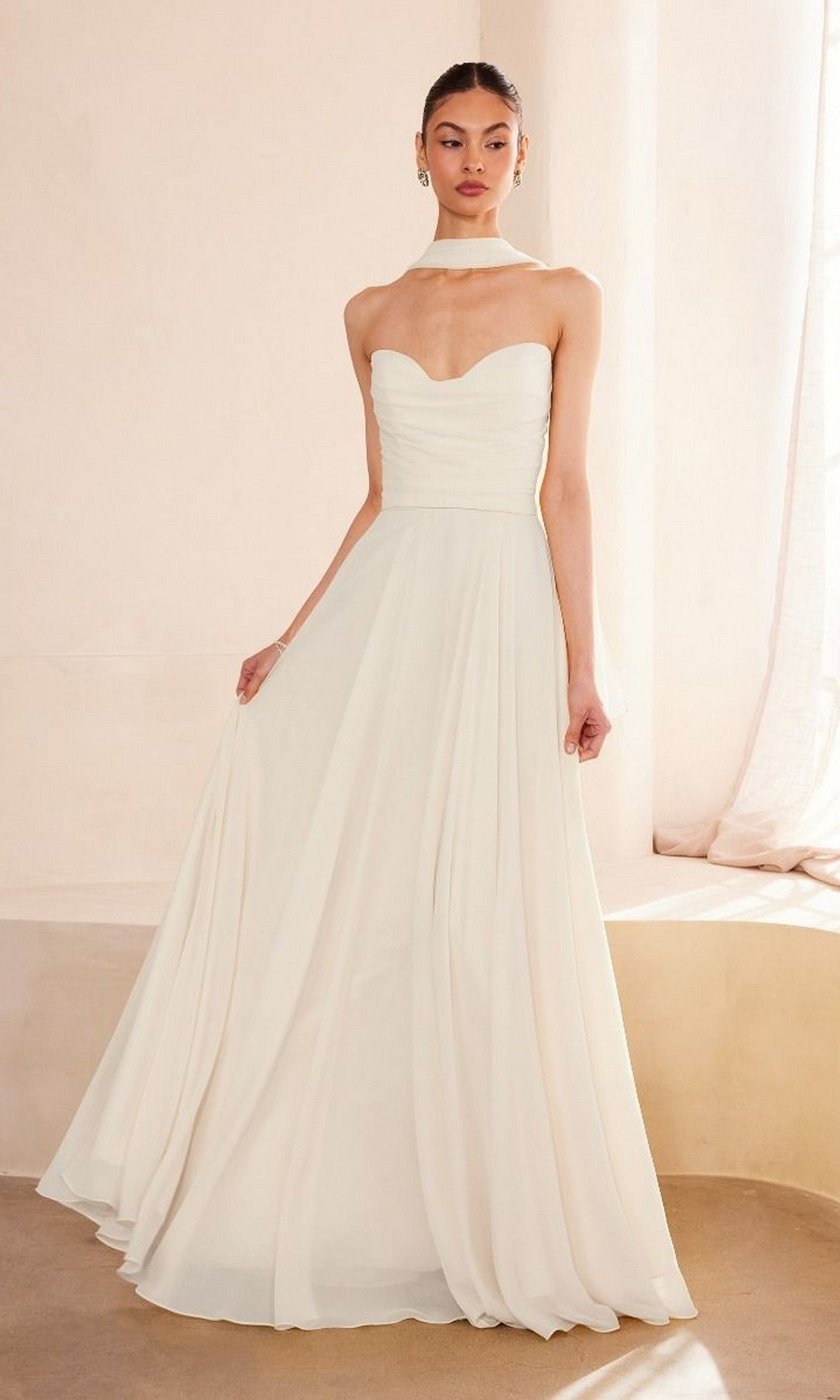 A woman indoors wears the Amoris T703W, a strapless long white wedding dress with a fitted bodice and flowing skirt, paired with a matching choker.